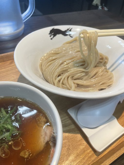 The image shows a bowl of noodles with a spoon inside, placed next to another bowl filled with soup. The bowls are sitting on a wooden table or tray. There is also a cup visible in the background. The main focus of the image is the noodle dish and its presentation, which includes the use of chopsticks for eating.