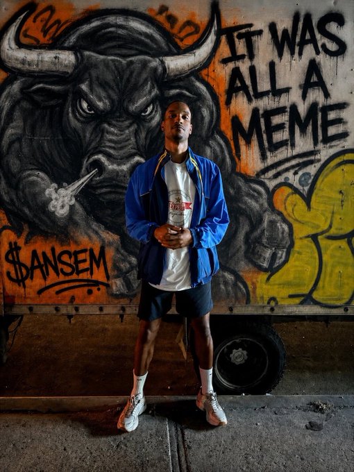 A young man in a blue jacket and shorts stands in front of a graffiti mural depicting a smoking bull with horns and the text 'IT WAS ALL A MEME' alongside '$ANSEM' and other tags.
