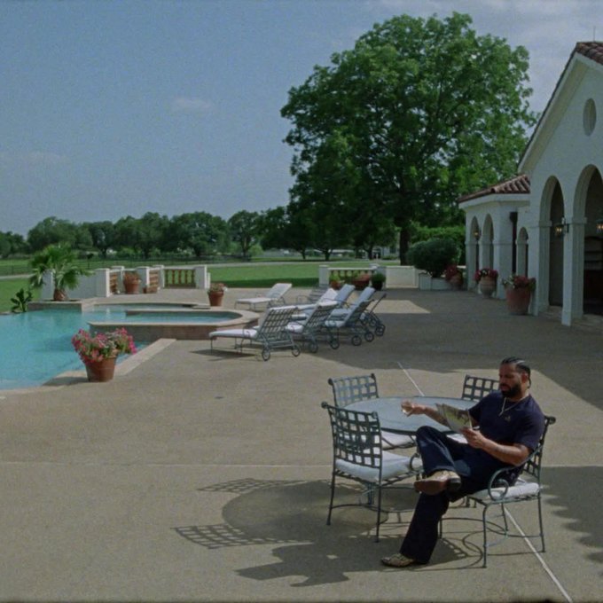 A scene depicting a bearded man relaxing at an outdoor patio beside a swimming pool at what appears to be a Spanish-style villa, reading a document or magazine.