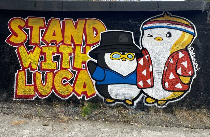 A colorful mural on a black wall reading 'STAND WITH LUCA' in yellow and red letters, featuring two cartoon penguin characters wearing a bucket hat and a headband.