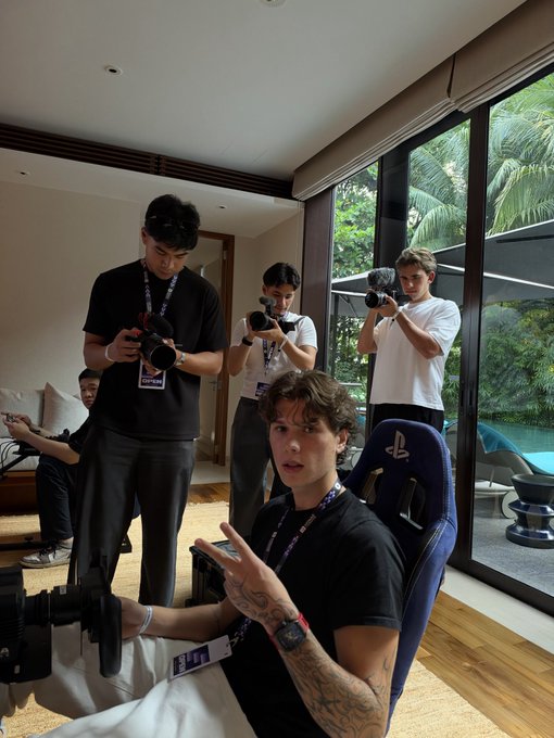 Several young men with cameras in a modern living room; one sits in a blue gaming chair in the foreground wearing lanyards.
