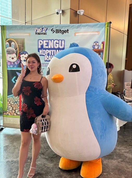 A woman in a black floral dress poses next to a large penguin mascot at a 'PENGU' event sponsored by Bitget.