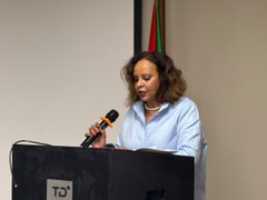 A woman in a light blue blouse speaks at a podium marked 'TGI' while reading from notes and speaking into a microphone.