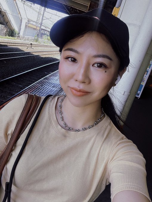 A selfie of a young woman wearing a black cap, beige ribbed top, and beaded necklace, taken on a train platform with railway tracks visible in the background.