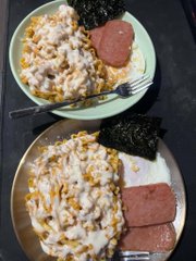 Two plates of food viewed from above, each containing white rice topped with corn and creamy sauce, sliced pink luncheon meat, a sheet of nori seaweed, and a fork.