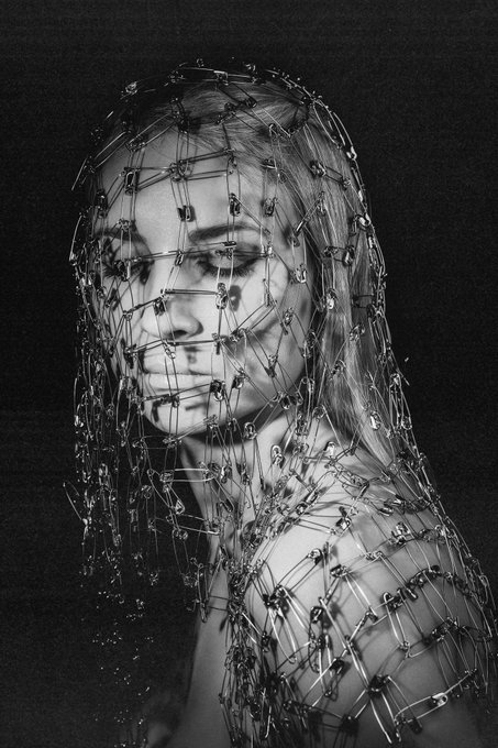 A black and white artistic portrait of a woman wearing a hood and body covering made entirely of linked safety pins against a dark background.