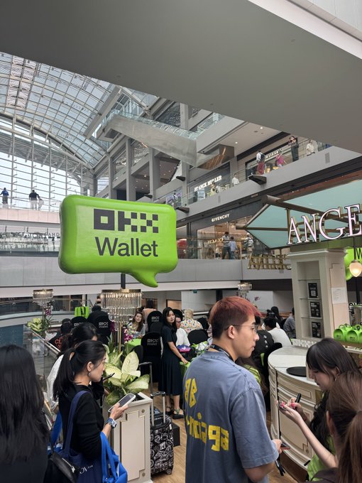 A photo taken inside a busy multi-level shopping mall, featuring a green 'OKX Wallet' speech bubble graphic overlaid on the scene with people browsing and walking below.