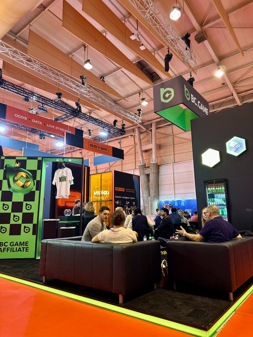A busy trade show or convention features a 'BC GAME AFFILIATE' booth with people sitting on dark leather couches in conversation.