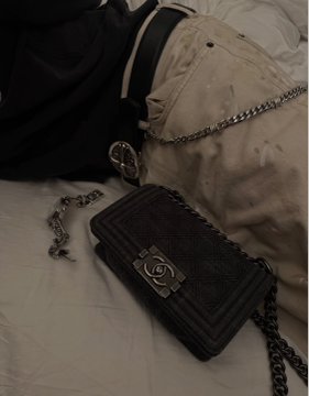 A black quilted Chanel handbag with a gold chain strap and interlocking CC turnlock clasp resting on white bedding beside light-colored pants.