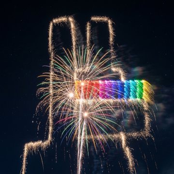 A fireworks display at night with a hand-shaped sparkle effect featuring a rainbow-colored palm in the center, set against a dark sky.