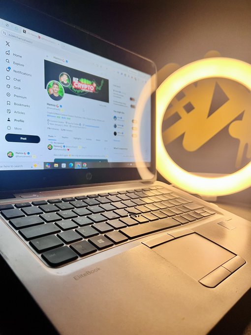 A photo of an HP EliteBook laptop displaying the X (Twitter) homepage with a cryptography-themed profile, positioned next to a glowing yellow Monero logo light.