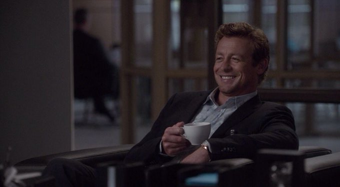 Simon Baker smiling while seated in a dark suit holding a white coffee cup.