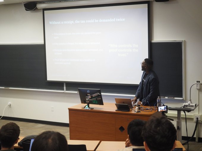 A speaker presents to graduate students in a classroom, with a projected slide titled "Without a receipt, the tax could be demanded twice" and the quote "Who controls the proof controls the trust," relating to XRPL Canada's lecture on XRP Ledger architecture and consensus mechanisms.