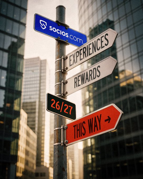 A street signpost displays signs reading 'socios.com', 'EXPERIENCES', 'REWARDS', '26/27', and a red 'THIS WAY →' arrow, set against a city skyline backdrop.