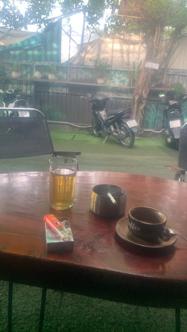 A wooden table holds a glass of beer, an ashtray with a cigarette, a coffee cup, and a pack of cigarettes, with motorcycles parked in the background.