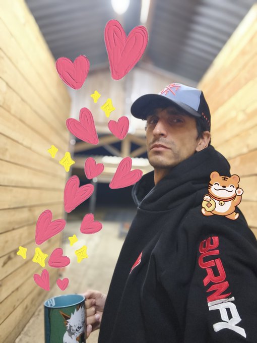 A selfie-style photo of a man wearing a black hoodie with 'BURN' written on the sleeve and a cap featuring the Union Jack, holding a mug while heart emojis and a lucky cat sticker are digitally overlaid onto the image.