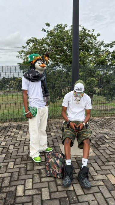 Two people pose on a brick-paved area, one wearing a penguin mask with a green cap and white outfit, while the other sits looking at a phone in a white t-shirt, camo shorts, and a green beanie, with a camo bag at their feet—matching the "GM CT Rise & Conquer" vibe with casual streetwear and playful energy.