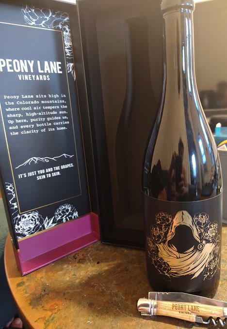 The image features a bottle of wine from Peony Lane Vineyards sitting next to its corkscrew. The wine bottle has a label with a picture of a person wearing a hood, which adds an interesting visual element to the scene. The corkscrew is placed on top of the wine bottle and appears to be in good condition. The image also includes text that reads "Finally opened my @PeonyLaneWine shipment from @scarcedotcity. This is how you ship something with taste. The box. The bottle. The little joy-sparker of a wine opener. Delivered with excellence and care, as only Bitcoiners know how to do."