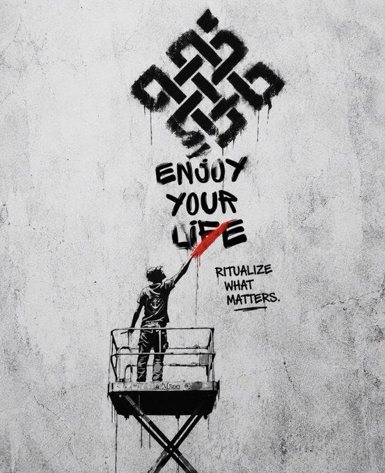 Street art by Smug on a concrete wall featuring a Celtic knot and a figure on a cherry picker painting the words 'ENJOY YOUR LIFE' and 'RITUALIZE WHAT MATTERS.'