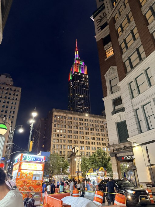 A nighttime street scene in New York City featuring the Empire State Building illuminated in rainbow colors, with street vendors, food carts, and crowds on the street below.