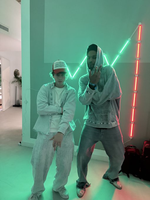 Two people pose in a neon-lit room with green and red LED tube lights forming geometric patterns on the wall behind them, both dressed in matching light-washed denim outfits with hoods and a trucker hat reading "UNCLE RICK'S AUTO SHOP." The "$ANSEM MODE" caption reflects the stylish, hypebeast aesthetic of the photo, suggesting the subjects are in a confident, trendy mood befitting the Ansem brand persona.