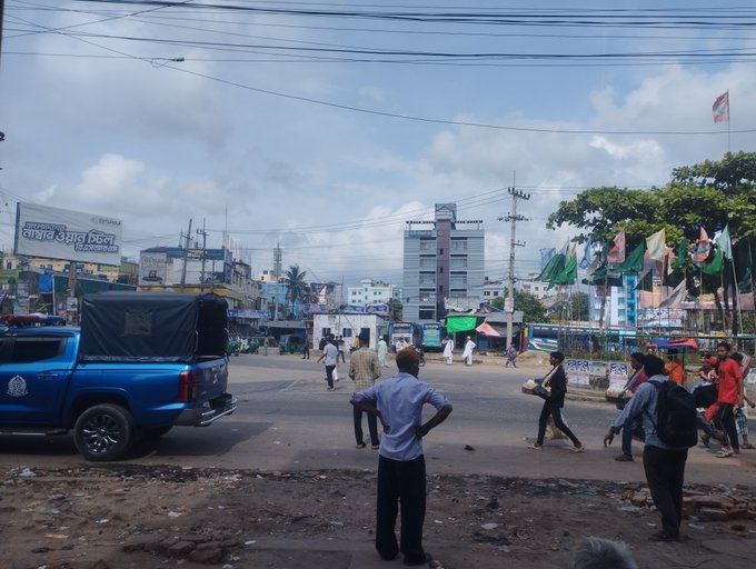 A bustling street scene in what appears to be a South Asian city, with a blue pickup truck parked on the left, people walking, buildings in the background, and signs in Bengali script visible on storefronts.