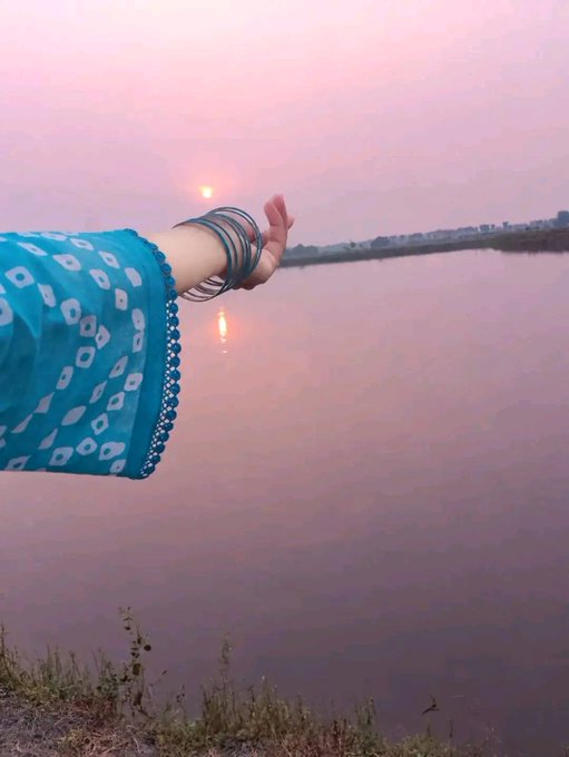 A person's arm wearing a turquoise bandhani-patterned sleeve and multiple bangles extends toward a sunset reflection on a calm lake, with distant buildings visible on the horizon.