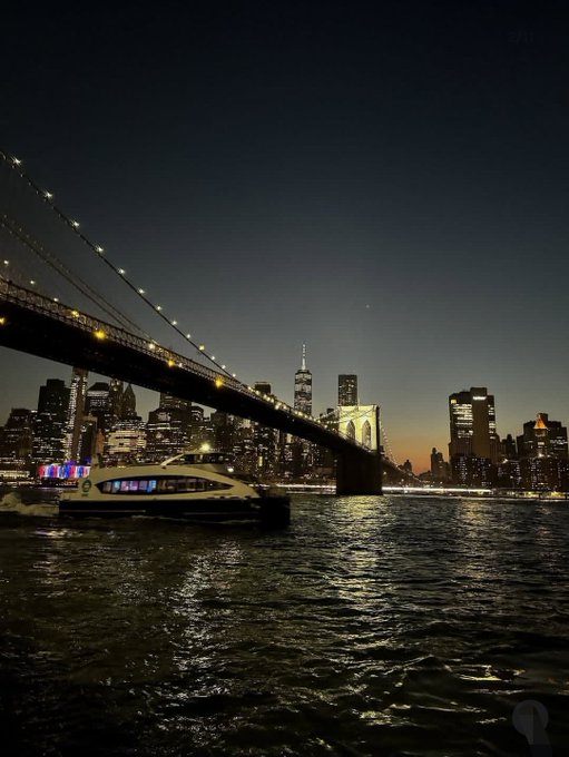 A nighttime photograph of the New York City skyline and the illuminated Brooklyn Bridge spanning the East River, with a lit boat passing underneath.