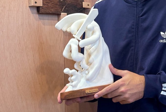 A person wearing an Adidas navy jacket holds a white sculptural piece mounted on a wooden base with a gold label reading 'To @venustwt'.