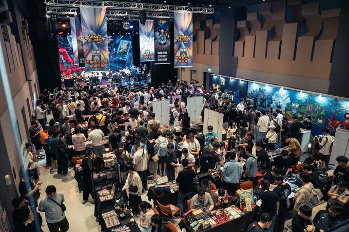 A crowded indoor event space with Pokémon-branded banners and signage, filled with attendees at various vendor tables.