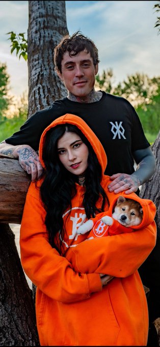 The image features a man wearing an orange hoodie and a woman holding a small dog in her arms. They are standing next to each other with their respective pets. The background of the photo includes trees, suggesting that they might be at a park or a similar outdoor location. The caption reads "My Bitcoin family @sautrah @thefocelot We just got the pictures back. Shout out to McGillis_Captures on instagram. Tons more from our family photo shoot coming soon. Bitcoin, Runestone, and $DOG go to the moon".