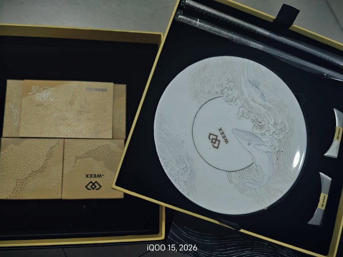 An open gold-themed gift box containing WEXX-branded tea boxes and an elegant white ceramic plate decorated with a whale design.