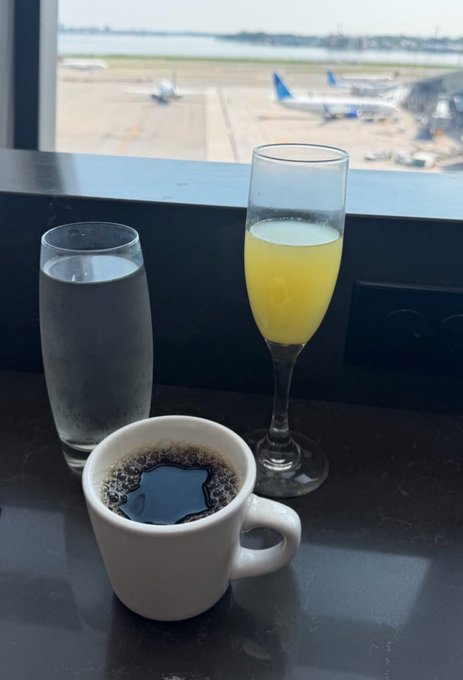 A black coffee mug, a glass of water, and a champagne flute of orange juice sit on a table overlooking an airport tarmac with planes visible through the window.
