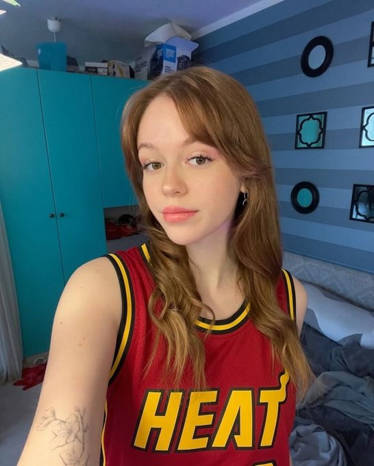 A young woman with long brown hair wearing a red and yellow 'HEAT' basketball jersey poses for a selfie in a room with teal cabinets and striped walls.