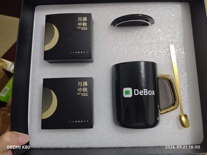 A DeBox gift set in a foam-lined box containing a black mug with gold handle, a gold spoon, and two boxes of Mid-Autumn Festival mooncakes (月满中秋), photographed with a Redmi K80 on 2026.09.21.
