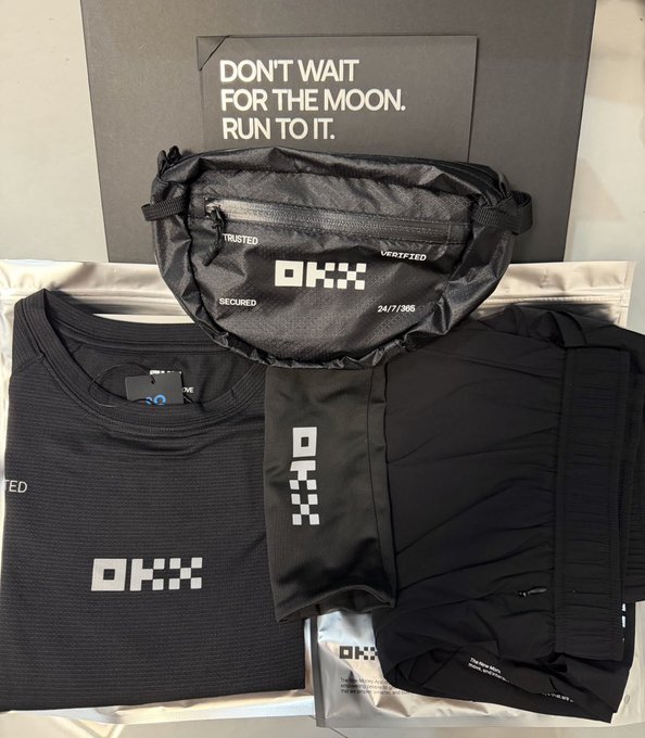 A collection of black OKX-branded apparel and accessories, including a fanny pack labeled 'TRUSTED' and 'SECURED', a t-shirt, and shorts, displayed in front of a box reading 'DON'T WAIT FOR THE MOON. RUN TO IT.'