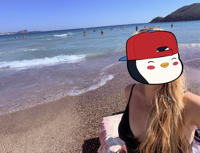 A woman in a black bikini on a beach with waves and mountains in the background, her face obscured by a cartoon emoji of a smiling character wearing a red cap.