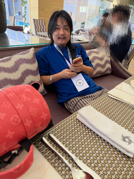 A person with long black hair wearing a blue t-shirt sits on a restaurant couch, looking at their phone, with a red Louis Vuitton backpack and table setting in the foreground.