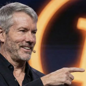 A close-up of a smiling gray-haired man in a dark shirt, gesturing with his finger while speaking, with an orange-lit blurred background.