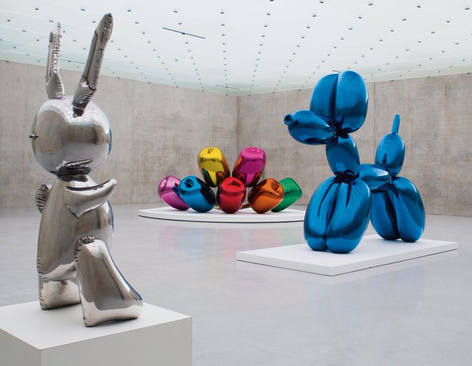 A Jeff Koons "Rabbit" sculpture and "Balloon Dog" sculptures are displayed in a gallery, prompting the post's tongue-in-cheek suggestion that Koons might be minting NFTs of similar balloon-themed artwork. The image showcases his iconic metallic and colorful balloon animal sculptures in a minimalist white space.