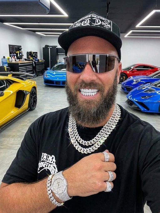 A man wearing a black cap, large sunglasses, a black t-shirt, and diamond-encrusted jewelry, including chains, rings, and a watch, smiling in a luxury garage filled with high-end sports cars.