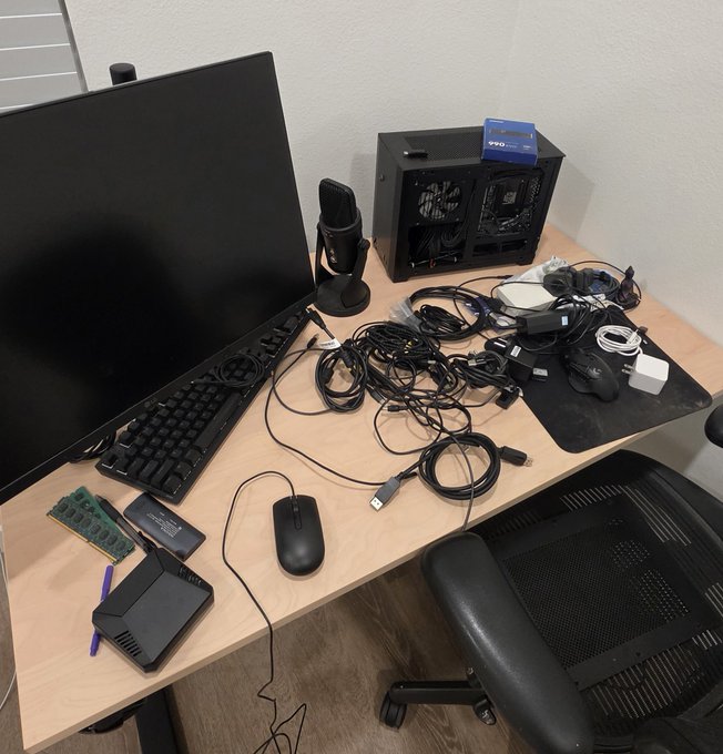 A wooden desk setup featuring a black monitor, mechanical keyboard, mouse, microphone, and an open computer case. The desk is cluttered with various cables, RAM, a pen, and peripherals, with an ergonomic black office chair tucked underneath.