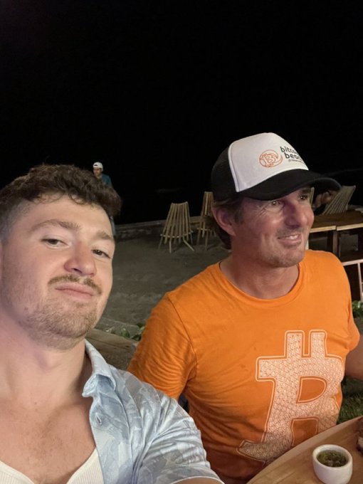 Two men pose for a selfie at an outdoor nighttime gathering, with the man on the right wearing an orange Bitcoin-themed t-shirt and a "little bit" cap. The post jokingly captions the scene as highlighting the man on the right's enthusiastic, "Tom Cruise-level" confidence in Bitcoin's bullish momentum.