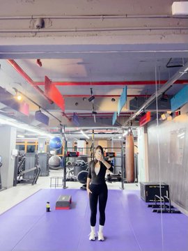 A woman stands in the middle of a brightly lit fitness studio with a purple floor, surrounded by gym equipment, exercise balls, and hanging colorful flags.