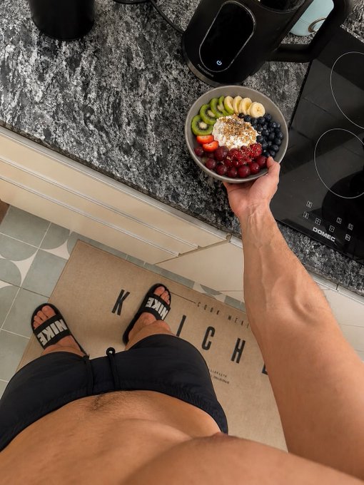 An overhead view of a shirtless person wearing black Nike slides holding a bowl of yogurt topped with sliced bananas, kiwi, strawberries, blueberries, cherries, and granola, while standing on a "KETCHUP" doormat in a kitchen.