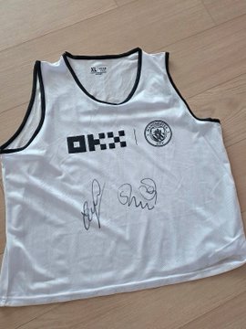 A white DKNY tank top with the number 5 and a circular emblem on the chest, featuring two signatures in black ink, lies on a wooden floor.