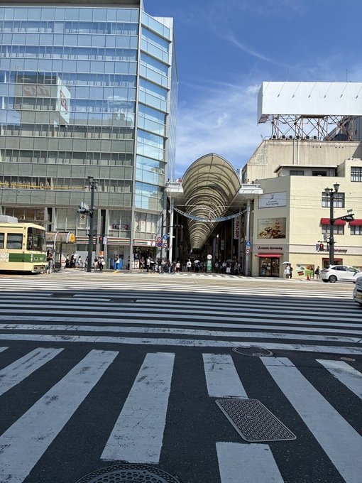 A sunny street view of a Japanese shopping arcade with a curved arcade roof, flanked by buildings, with a vintage tram on the left and a wide pedestrian crosswalk in the foreground.