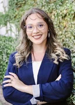 A portrait of N'Kiyla "Jasmine" Thomas, a Democratic Socialist who has won Oklahoma's Democratic primary for U.S. Senate according to Polymarket projections, smiling with arms crossed in front of a leafy green background.