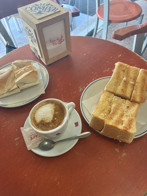 A cafe table with a cappuccino on a saucer, a toasted sandwich cut into pieces on a plate, and a pastry on another plate, with a wooden 'Serra COMPAL' menu/napkin holder.