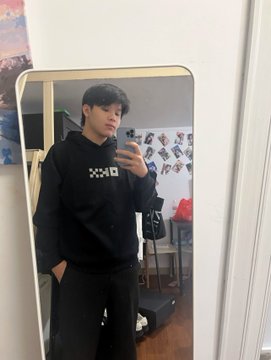A mirror selfie of a young man wearing a black hoodie with a checkered logo, standing in a room with anime/manga posters on the wall behind him.